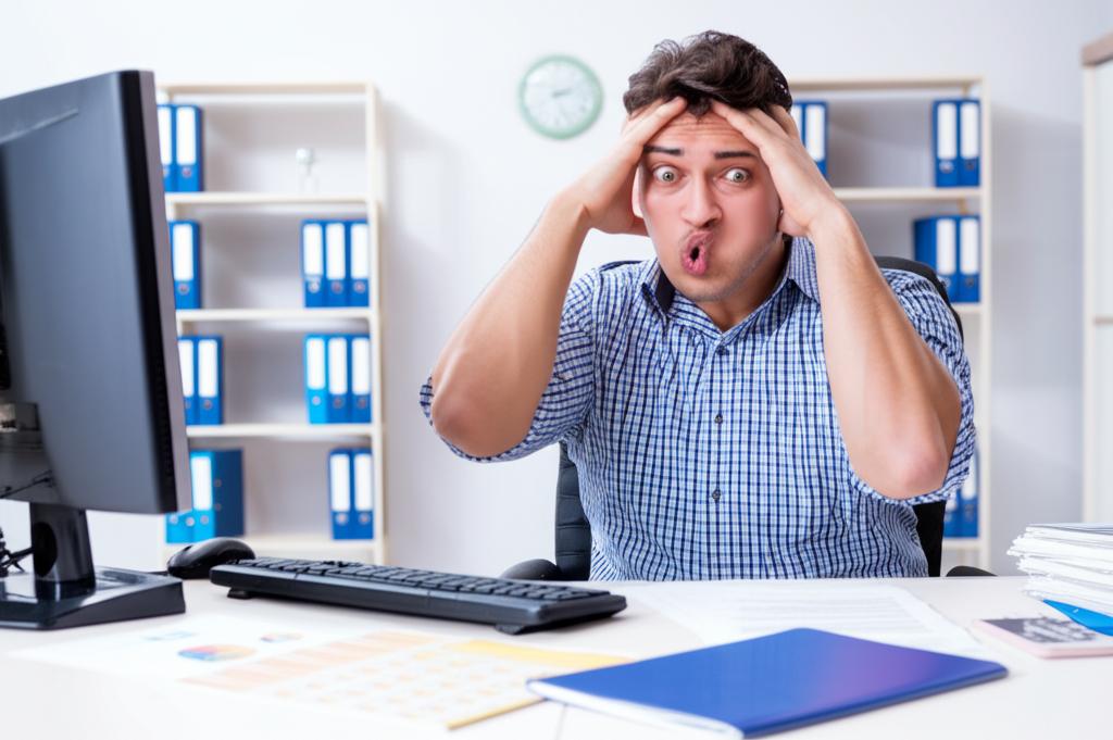 man sitting at desk looking stressed and tired, surrounded by paperwork and computer screens, low energy, office environment