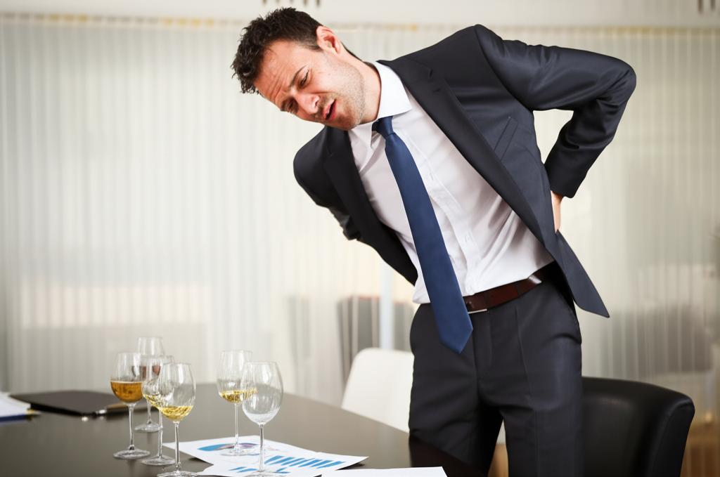 Image prompt: businessman in a suit looking tired and holding his lower back, surrounded by wine glasses and business documents